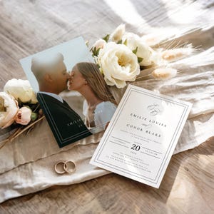 Puede incluir: Conjunto de papelería de boda con una tarjeta fotográfica de una pareja besándose, una invitación formal con texto y dos anillos de boda dorados. Flores blancas y vegetación dispuestas alrededor de la papelería sobre una superficie neutra.