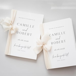 May include: Two white wedding programs with ivory ribbon and bow accents. The programs display the names "CAMILLE and ROBERT", the date "26.06.2026", and the location "frise bonaparte hotel, PARIS FRANCE".