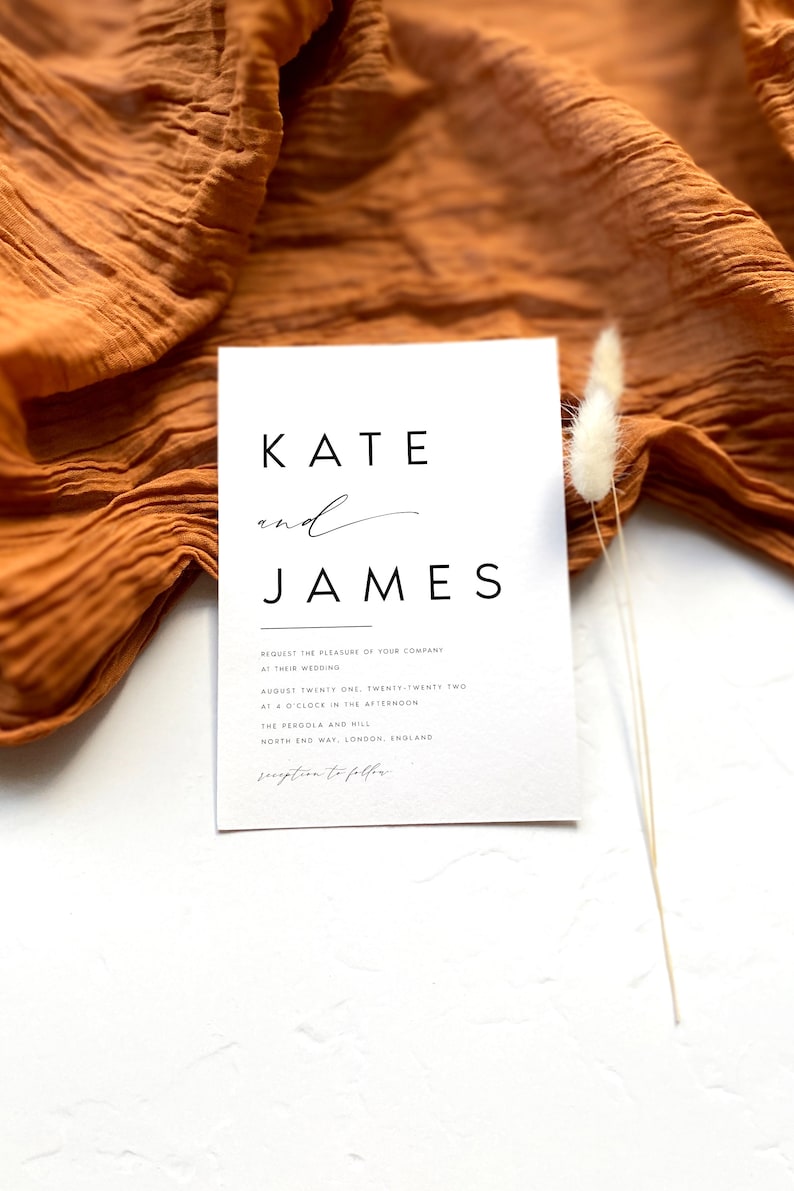 May include: A minimalist wedding invitation in white, with the names "Kate and James" in black. The invitation includes event details and is set against a textured orange fabric and a white background, with a sprig of dried flowers.