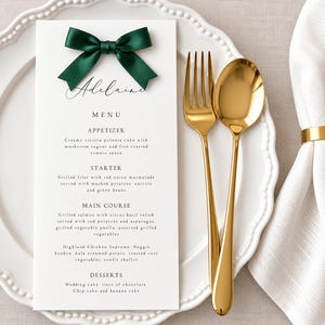 May include: A white rectangular menu card with the name Adeline in black script, topped with a dark green satin bow. The menu lists courses including appetizer, starter, main course, and desserts. A gold fork and spoon rest on a white plate.
