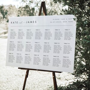 May include: A white wedding seating chart on a wooden easel. The chart lists names under table numbers. The top of the chart reads "KATE and JAMES" and "PLEASE TAKE A SEAT 24.07.2022. NEW YORK."