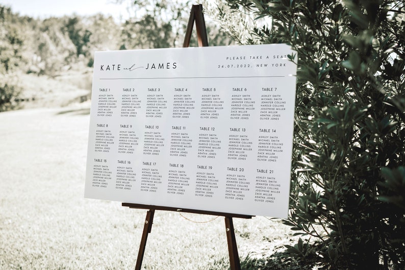 May include: A white wedding seating chart on a wooden easel. The chart lists names under table numbers. The top of the chart reads "KATE and JAMES" and "PLEASE TAKE A SEAT 24.07.2022. NEW YORK."