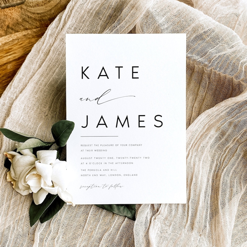 May include: A minimalist wedding invitation in white with black text. The names "KATE" and "JAMES" are prominently displayed. The invitation includes the wedding date, time, and location details. A white flower and green leaves are in the lower left corner. The invitation is on beige fabric.
