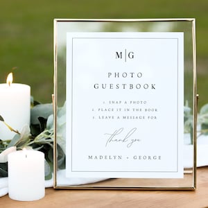 May include: A gold-framed sign with white paper inside. The sign says "MG Photo Guestbook" and includes instructions for guests to take a photo, place it in the book, and leave a message. The sign also says "Thank you Madelyn + George".