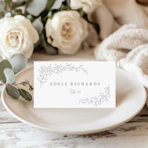 May include: A white place card with the name "ADELE RICHARDS" and "Table 10" printed in gray. The card is decorated with a gray floral design and sits on a white plate. Soft white roses and greenery are in the background.