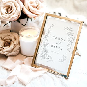 May include: A gold-framed sign with a white background and black calligraphy that reads "Cards and Gifts Thank You". The sign is decorated with a simple floral wreath design. The sign is sitting on a white surface with a candle and roses.