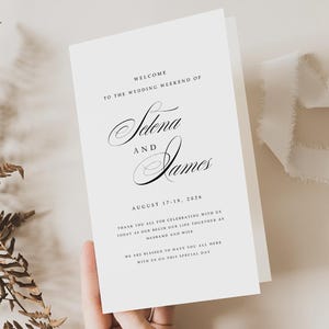 Könnte beinhalten: Eine weiße Hochzeits-Willkommenskarte mit eleganter Schrift. Die Karte lautet: "Willkommen zum Hochzeitswochenende von Selena und James, 17.-19. August 2026." Zusätzlicher Text drückt Dankbarkeit gegenüber den Gästen aus.