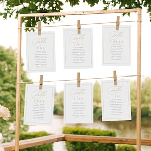 May include: Six white cards with gold trim and calligraphy are hung on a gold metal stand. Each card has a table number and a list of names. The cards are labeled Table One, Table Two, Table Three, Table Four, Table Five, and Table Six.