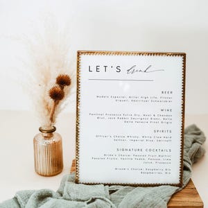 May include: A framed menu with the text "LET'S Drink" in elegant script. The menu lists beer, wine, spirits, and signature cocktails. A small vase with dried flowers and a green cloth are in the foreground.