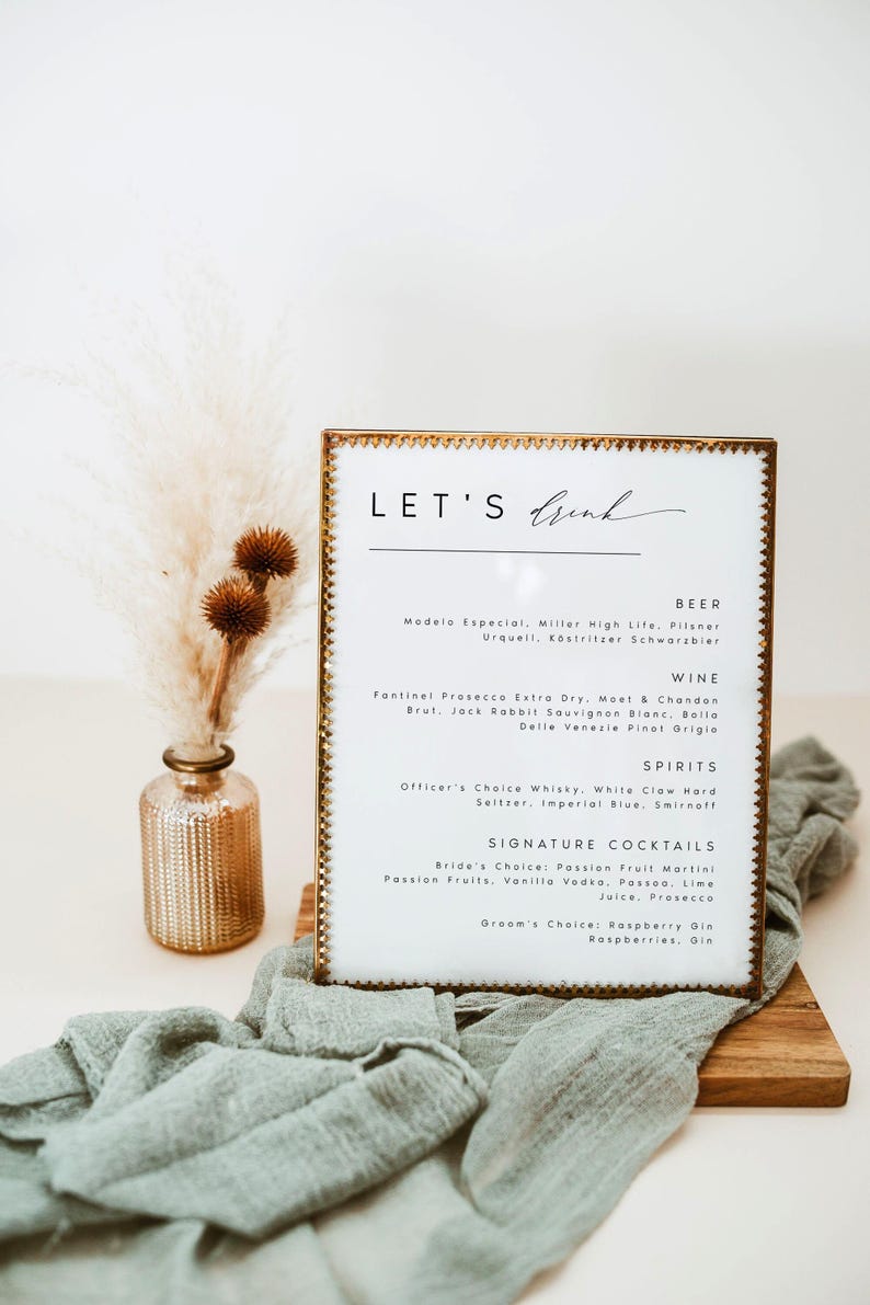 May include: A framed menu with the text "LET'S Drink" in elegant script. The menu lists beer, wine, spirits, and signature cocktails. A small vase with dried flowers and a green cloth are in the foreground.