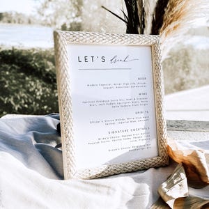 May include: A framed menu with the text "LET'S Drink" at the top. The menu lists beer, wine, spirits, and signature cocktails. The frame has a textured, light-colored design, and the background is blurred.