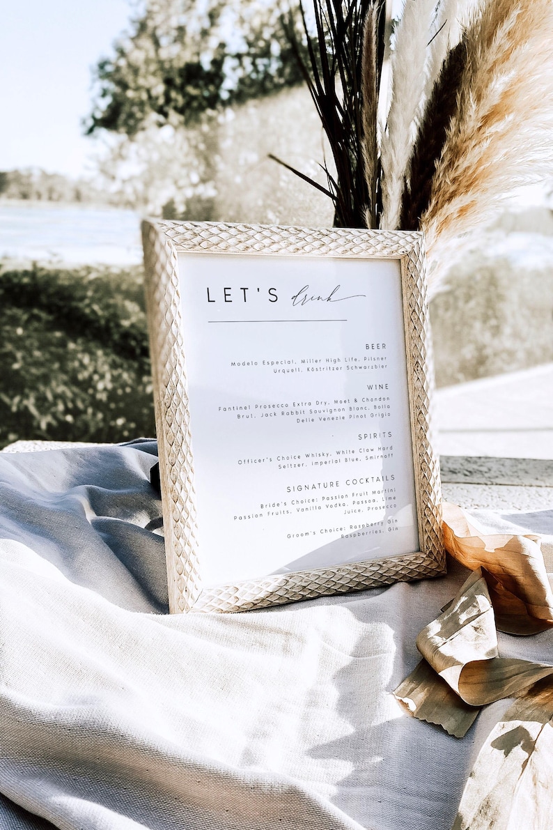 May include: A framed menu with the text "LET'S Drink" at the top. The menu lists beer, wine, spirits, and signature cocktails. The frame has a textured, light-colored design, and the background is blurred.