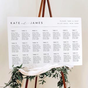 May include: A white rectangular wedding seating chart sign with black text reading Kate and James, Please Take a Seat, 24.07.2022, New York, and a list of tables 1 through 21 with guest names. The sign is displayed on a wooden easel with greenery.