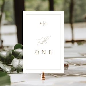 May include: White table number card with a gold border and gold text. The card reads "M|G Table ONE". The card is on a gold stand.