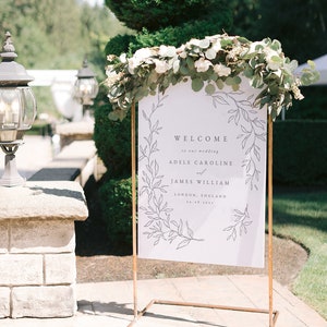 May include: A white welcome sign with a floral design and the text "Welcome to our wedding Adele Caroline and James William London, England 24.08.2023". The sign is on a copper stand and decorated with greenery.