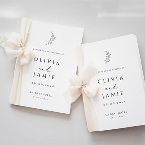 May include: Two white wedding programmes featuring black text that reads WELCOME TO THE WEDDING OF OLIVIA and JAMIE 26.06.2026 LA REVE HOTEL PARIS FRANCE. Each programme is adorned with a delicate black line drawing of a leafy branch and a cream ribbon bow.