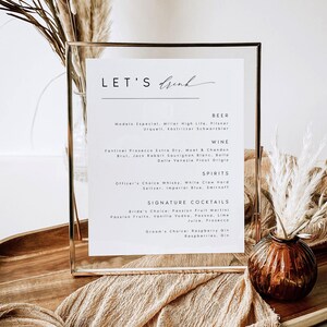 May include: A framed menu with the words "LET'S Drink" in elegant script. The menu lists beer, wine, spirits, and signature cocktails. The frame is gold, and the menu is on a wooden tray with decorative elements.