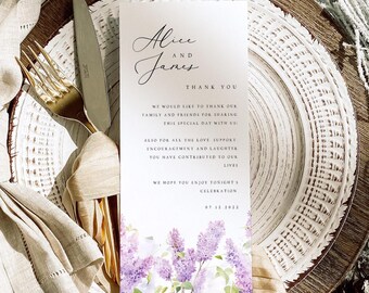 May include: A wedding place card with the names "Alice and James" and a thank you message. The card features a watercolor floral design in shades of purple and green. The card is placed on a decorative plate with a gold fork and knife.