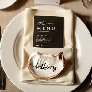 May include: A white oyster shell with a gold rim, used as a place card holder. The name "anthony" is written in black calligraphy on the inside of the shell.