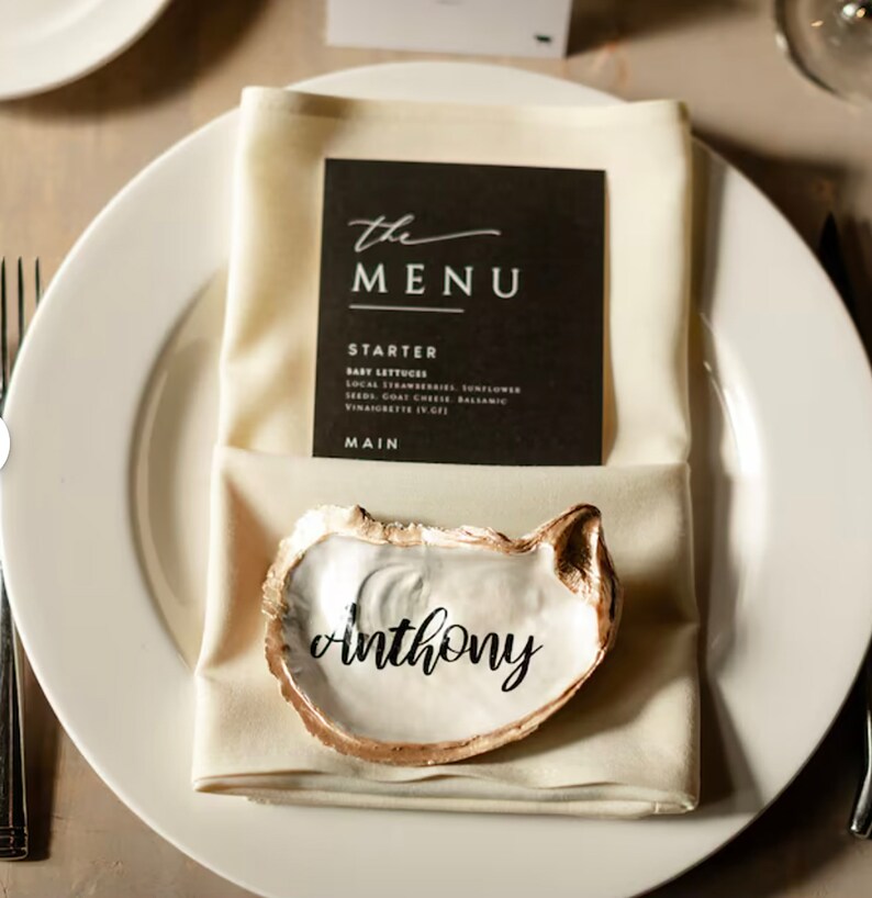 May include: A white oyster shell with a gold rim, used as a place card holder. The name "anthony" is written in black calligraphy on the inside of the shell.