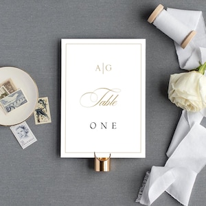 May include: White table number card with gold trim and the text "A|G Table ONE" in black font.