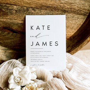 May include: A white wedding invitation with the names "KATE" and "JAMES" in a minimalist font. The invitation includes the wedding details: date, time, and location. The invitation is placed on a white fabric with white flowers.