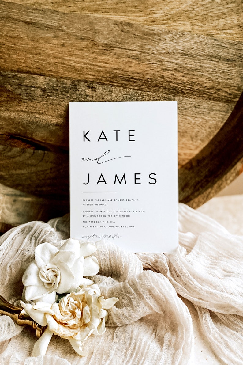 May include: A white wedding invitation with the names "KATE" and "JAMES" in a minimalist font. The invitation includes the wedding details: date, time, and location. The invitation is placed on a white fabric with white flowers.