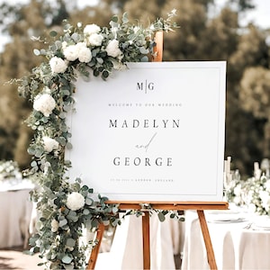 May include: White wedding sign with black calligraphy text. The sign reads "M/G Welcome to our wedding Madelyn and George 18.08.2021 London England". The sign is propped up on an easel with greenery and white flowers.