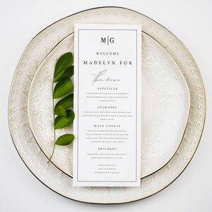May include: A wedding menu card with the text "MG WELCOME MADELYN FOX" in a minimalist design. The menu card is placed on a white plate with a gold rim, and a sprig of green leaves is placed on the plate.