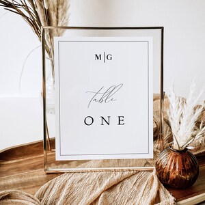 May include: A gold-framed table number sign with the text "M|G Table ONE" on a white background. The sign is sitting on a wooden tray with a beige fabric runner and a small vase with dried pampas grass.