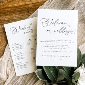 Pode incluir: Dois cartões de papelaria de casamento brancos com escrita preta elegante. Um cartão diz "Bem-vindo ao nosso casamento" e o outro lista "Eventos de fim de semana" com um cronograma. Os cartões estão em uma superfície de madeira com vegetação.