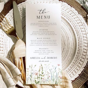 Könnte beinhalten: Eine Hochzeitsmenükarte mit floralen Illustrationen am unteren Rand, mit den Namen Amelia und Robert und dem Datum 26.06.2026. Das Menü listet Vorspeisen, Vorspeisen, Hauptgerichte und Desserts auf. Die Karte liegt auf einem weißen Teller mit einer goldenen Gabel und einem Messer.