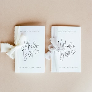Peut inclure: Deux programmes de mariage blancs avec du texte noir. Les programmes sont li&eacute;s avec un ruban blanc. Le texte indique "Bienvenue au mariage de Nathalie Tyler" et comprend la date et le lieu du mariage.