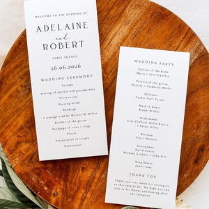 May include: Two white wedding ceremony programs with black text, one listing the ceremony details and the other the wedding party. The programs are on a wooden tray with greenery. The ceremony is in Paris, France.