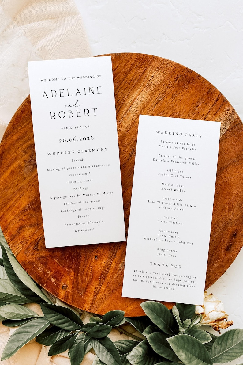 May include: Two white wedding ceremony programs with black text, one listing the ceremony details and the other the wedding party. The programs are on a wooden tray with greenery. The ceremony is in Paris, France.