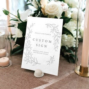 Puede incluir: Un cartel blanco con un borde floral y el texto "FULLY EDITABLE CUSTOM SIGN DESIGN AND PRINT UNLIMITED SIGNS". El cartel está sobre una mesa con un mantel marrón.