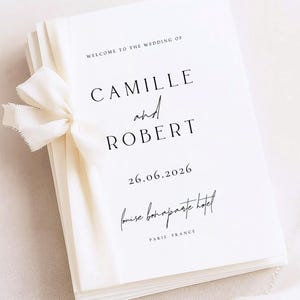 Könnte beinhalten: Ein Stapel weißer Hochzeitskarten, gebunden mit einem cremefarbenen Band. Die Karten zeigen schwarzen Text: "Welcome to the Wedding of Camille and Robert", das Datum "26.06.2026" und den Veranstaltungsort "louise bonaparte hotel" in Paris, Frankreich. Das Band ist gebunden.