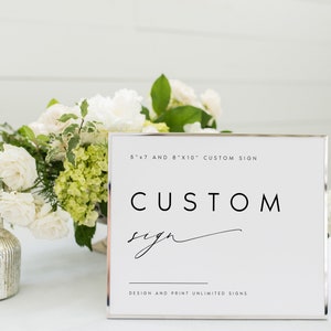 May include: A white sign with a silver frame that says "CUSTOM sign" in black text. The sign is sitting on a table with white flowers and greenery. The text at the top of the sign says "5"x"7" and 8"x"10" CUSTOM SIGN. The text at the bottom of the sign says "DESIGN AND PRINT UNLIMITED SIGNS."