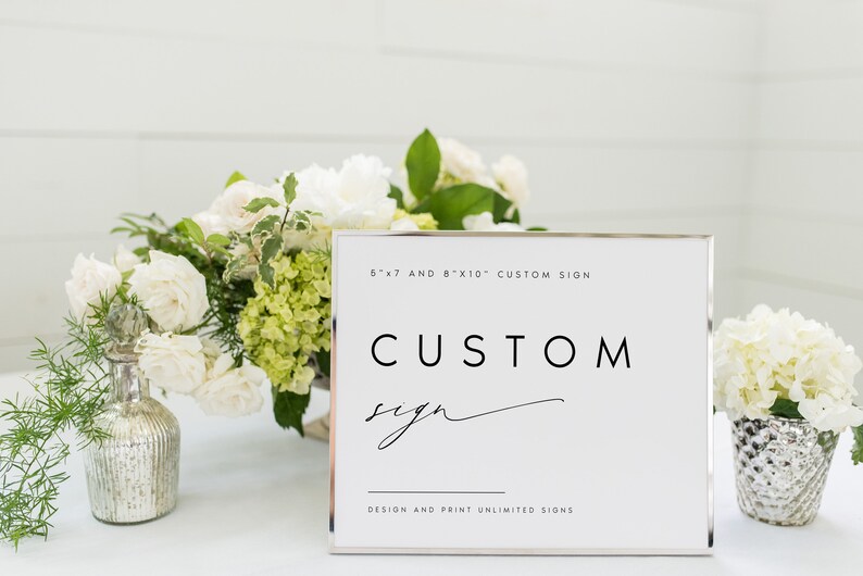 May include: A white sign with a silver frame that says "CUSTOM sign" in black text. The sign is sitting on a table with white flowers and greenery. The text at the top of the sign says "5"x"7" and 8"x"10" CUSTOM SIGN. The text at the bottom of the sign says "DESIGN AND PRINT UNLIMITED SIGNS."