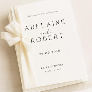 May include: A stack of white wedding programs tied with a cream-colored ribbon. The programs feature the names "Adelaine and Robert" and the date "26.06.2026". The venue is listed as "La Reve Hotel, Paris, France".