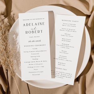 May include: Two elegant wedding ceremony programs on a white plate, featuring the names "Adelaine and Robert" and the date "26.06.2026". The program details the ceremony and wedding party. A dried plant stem adds a natural touch.