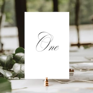 May include: White card with the word "One" written in cursive script on a gold metal stand.