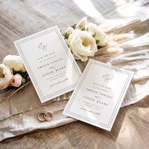 May include: Two white wedding invitations with black text, featuring the names Emilie Louisa and Conor Blake, and gold wedding rings. The invitations are surrounded by white roses and other flowers.