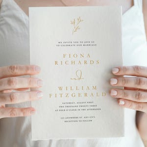 May include: A wedding invitation with gold lettering on a cream-colored card. The text includes the names Fiona Richards and William Fitzgerald, along with the wedding date and time. A floral design is at the top.