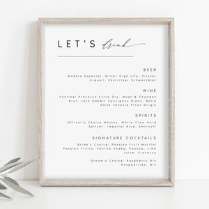 May include: A framed print with the words "LET'S Drink" in elegant script. The print lists beer, wine, spirits, and signature cocktails, including Modelo Especial, Moet & Chandon, and Passion Fruit Martini. The frame is a light wood color.