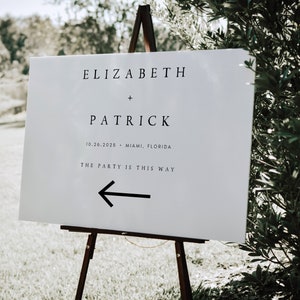 May include: White wedding sign with black text that reads "Elizabeth + Patrick 10.26.2026 - Miami, Florida The Party is This Way" with a black arrow pointing left.
