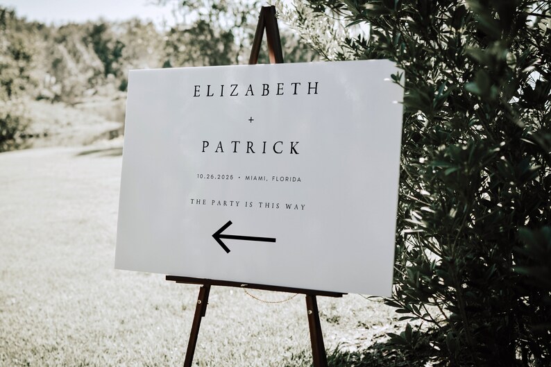 May include: White wedding sign with black text that reads "Elizabeth + Patrick 10.26.2026 - Miami, Florida The Party is This Way" with a black arrow pointing left.