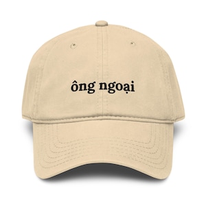 Puede incluir: Gorra de béisbol beige con el texto vietnamita "ông ngoại" bordado en la parte delantera. La gorra tiene una visera curva y una correa ajustable en la parte trasera. El sombrero está hecho de una tela suave y duradera.