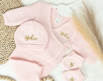 May include: A baby clothing set in soft pink, including a knitted wrap-style cardigan, a beanie, and two pairs of mittens. The name "Julia" is embroidered in gold on the hat and cardigan. The set is displayed on a white hanger.