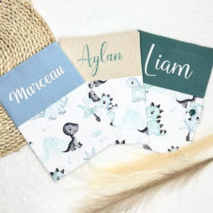 Peut inclure: Trois couvertures pour bébés avec des noms personnalisés. Chaque couverture présente un imprimé de dinosaures blancs avec des accents bleus et gris. Les sections supérieures sont de couleurs unies : bleu clair, beige et vert d'eau. Les noms "Marceau", "Aylan" et "Liam" sont écrits sur chaque couverture.
